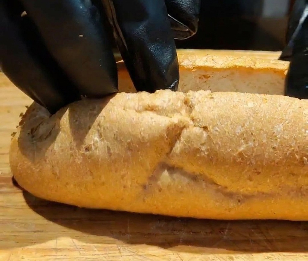 Close-up of hands in black gloves pulling open a sliced whole wheat baguette on a wooden board to create a space for filling.