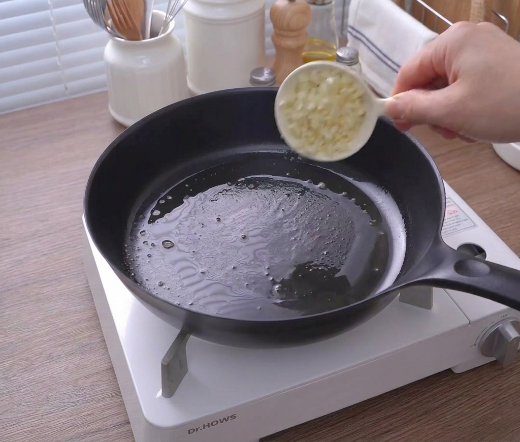 A small white cup pouring minced garlic into a black skillet containing melted butter.