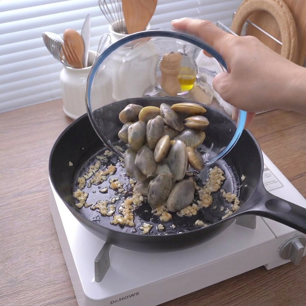A glass bowl pouring fresh, closed clams into a black skillet containing sautéed minced garlic.