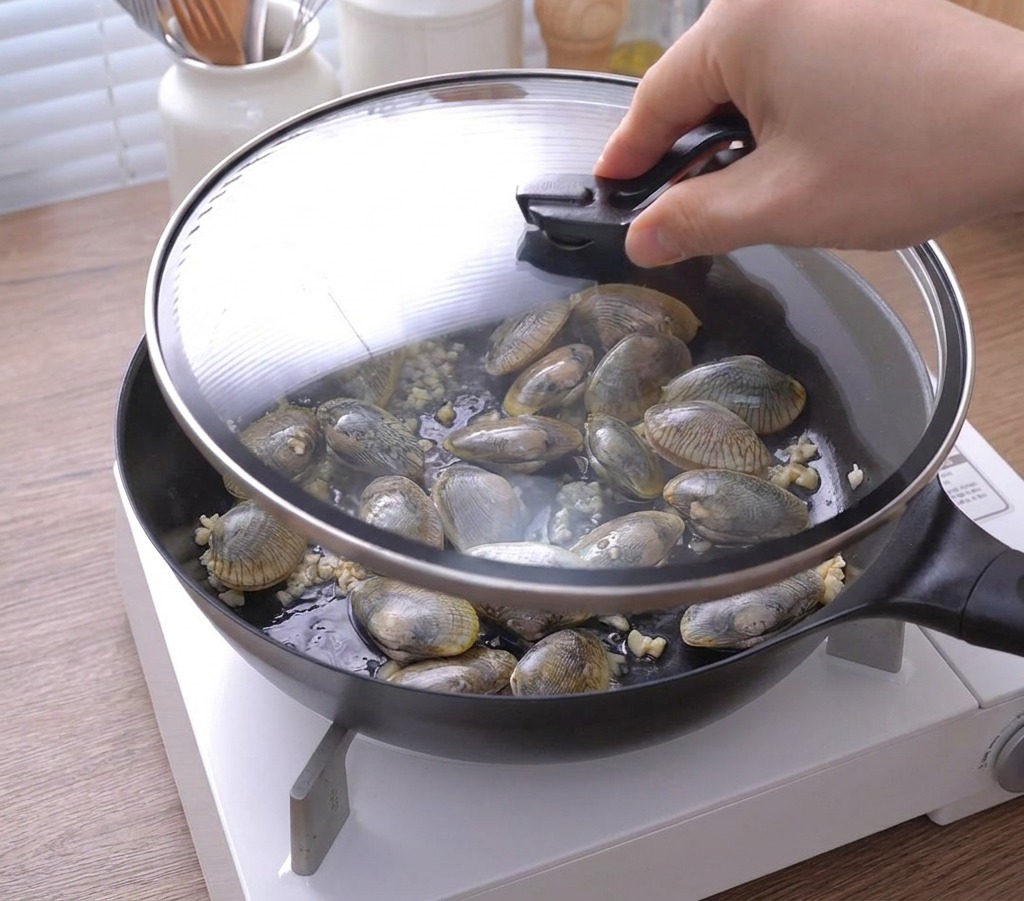 A hand placing a glass lid over a skillet filled with clams and sautéed garlic.