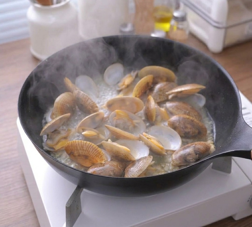 Steam rising from a black skillet filled with many fully opened and cooked clams.