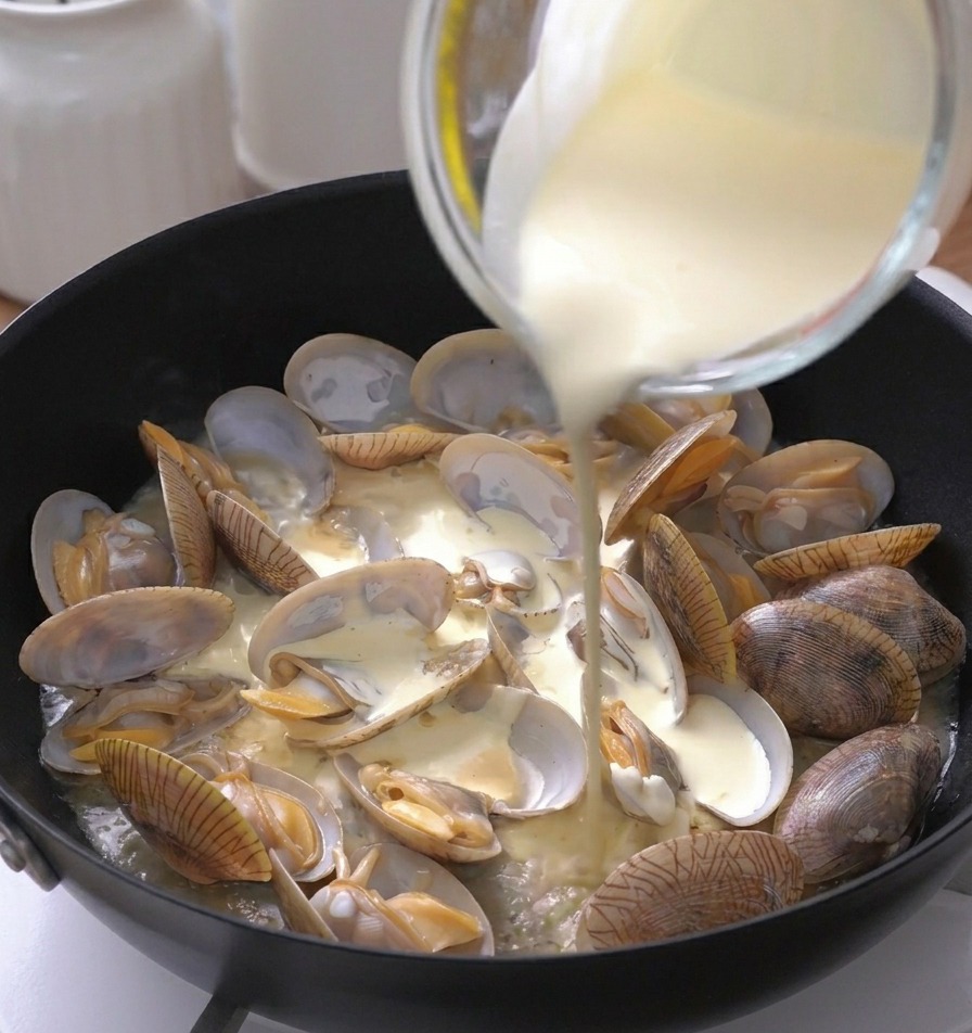 Thick white heavy cream being poured from a glass measuring cup into a pan filled with cooked, open clams.