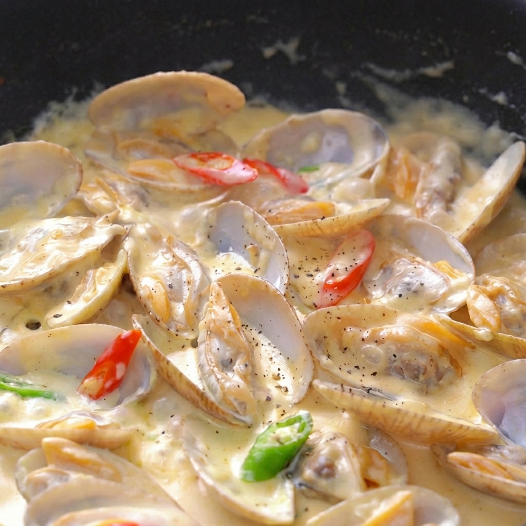 A close-up view of cooked clams in a thick, bubbling cream sauce, topped with black pepper, parsley flakes, and chili slices.