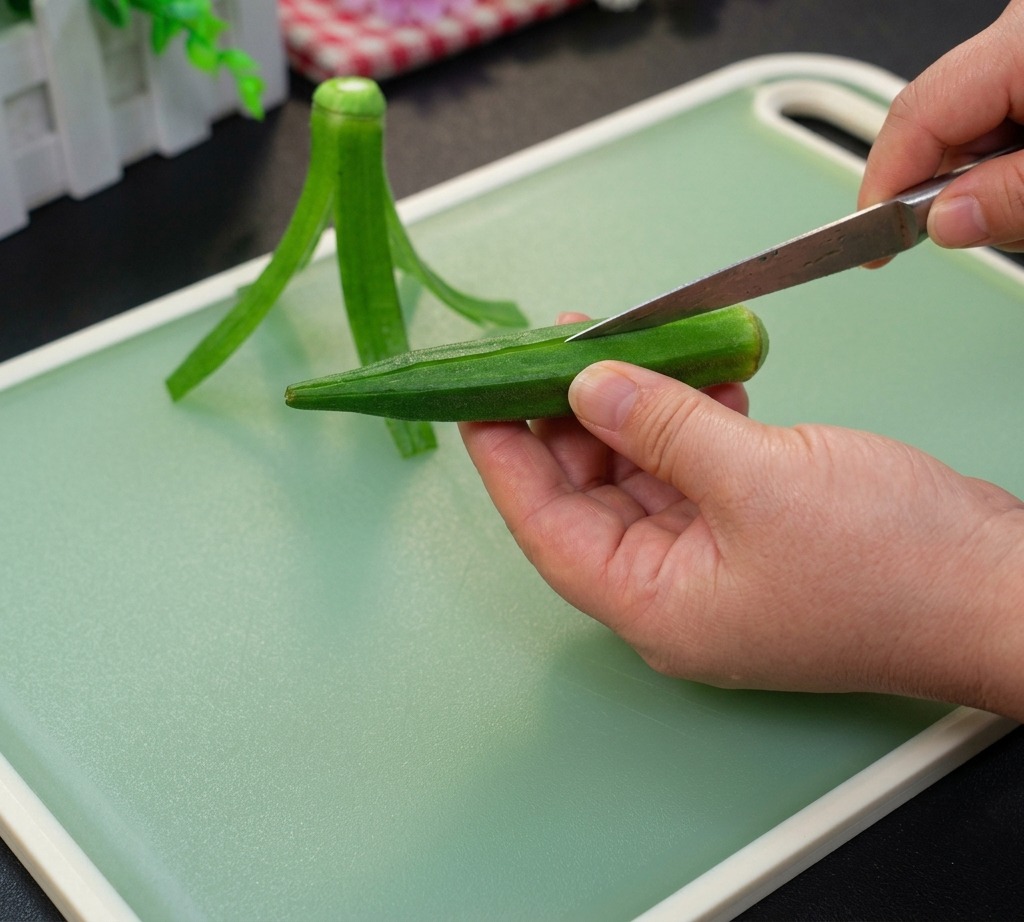 A hand holding a whole green okra pod on a cutting board while a knife starts to cut the top edge.