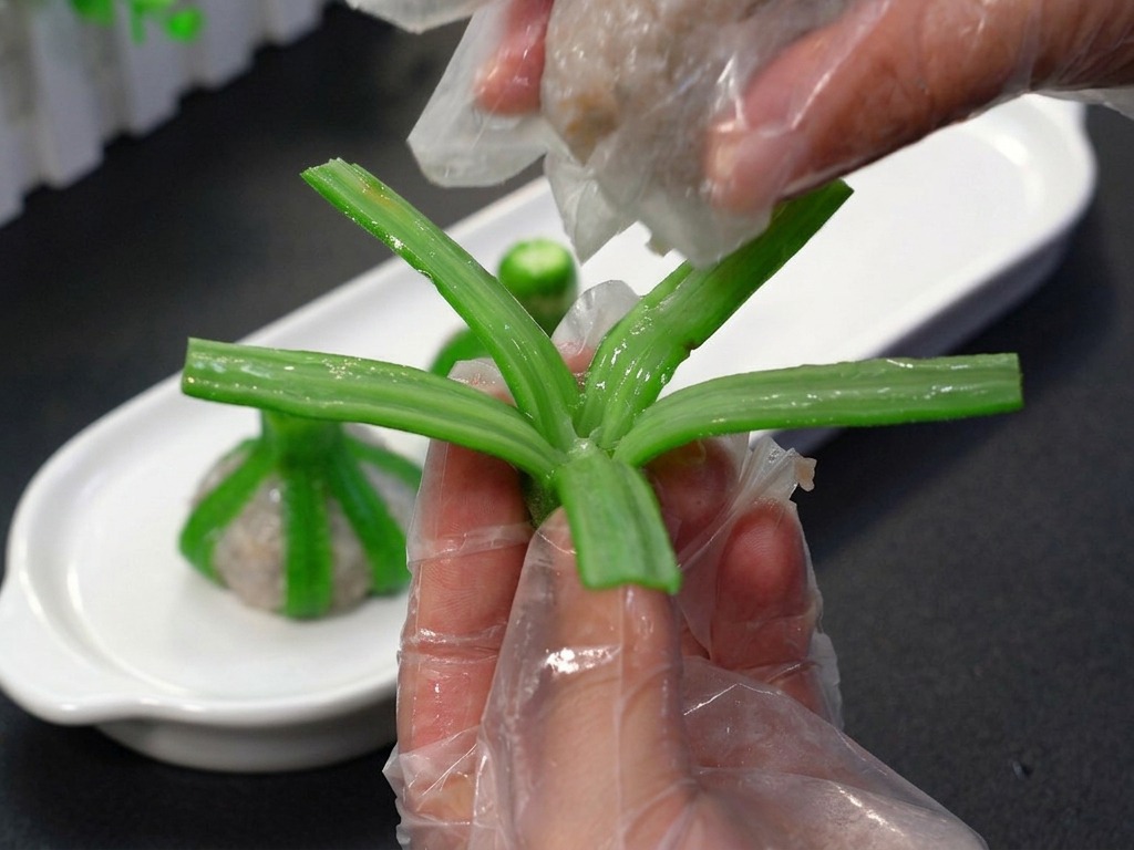 A person in plastic gloves placing a ball of pinkish shrimp mince into the center of blanched okra strips.