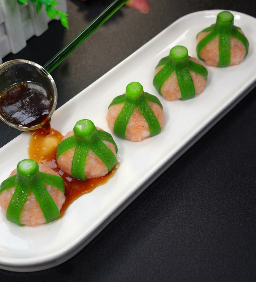 A close-up of a hand pouring dark savory sauce from a small glass pitcher onto five steamed, shrimp-stuffed okra pieces arranged on a white serving plate.