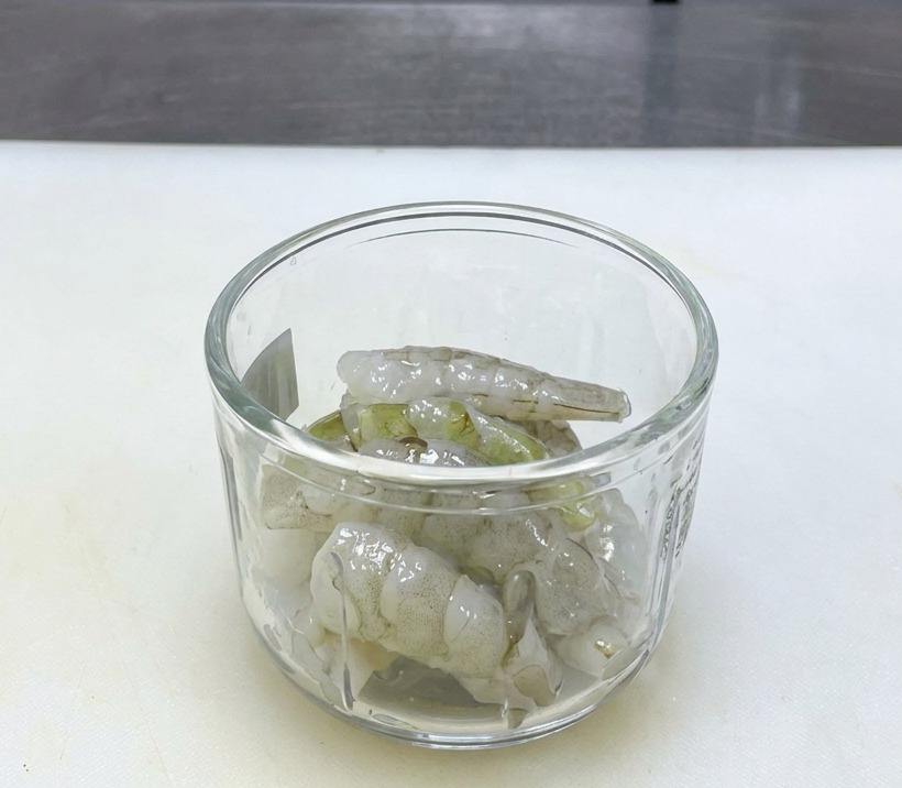 Raw translucent shrimp sitting in a clear glass blender bowl on a clean white cutting board.