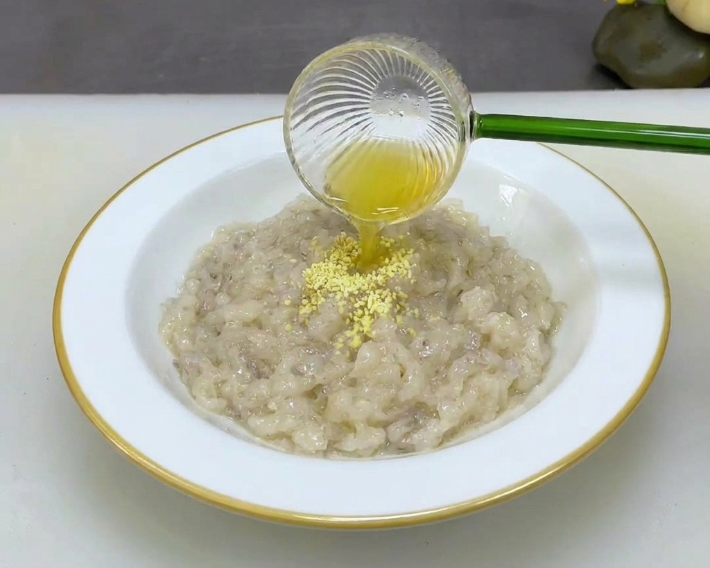 A person pouring fine yellow seasoning powder from a small glass scoop into a white bowl filled with thick, mashed shrimp paste.