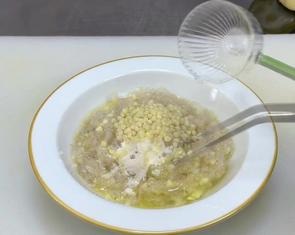 Metal tweezers stirring small round cream colored okra seeds into a pale thick shrimp paste mixture within a white bowl.