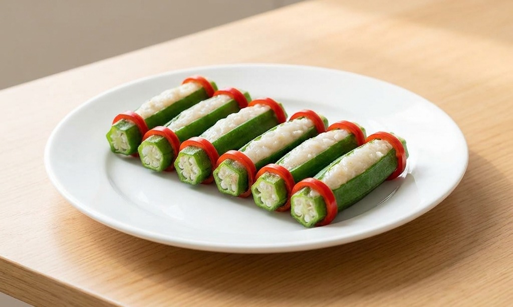 A white plate featuring a neat row of green okra pods stuffed with shrimp paste, each garnished with two bright red chili rings.