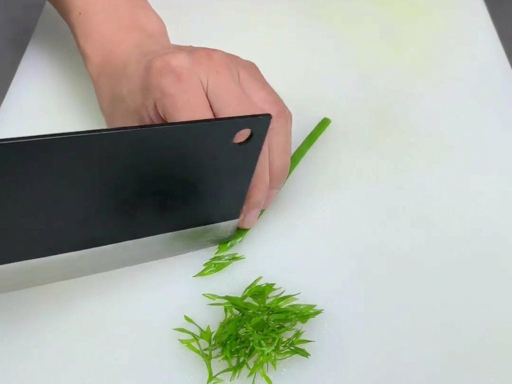 A person using a large black cleaver to finely shred green onion stalks on a white cutting board.