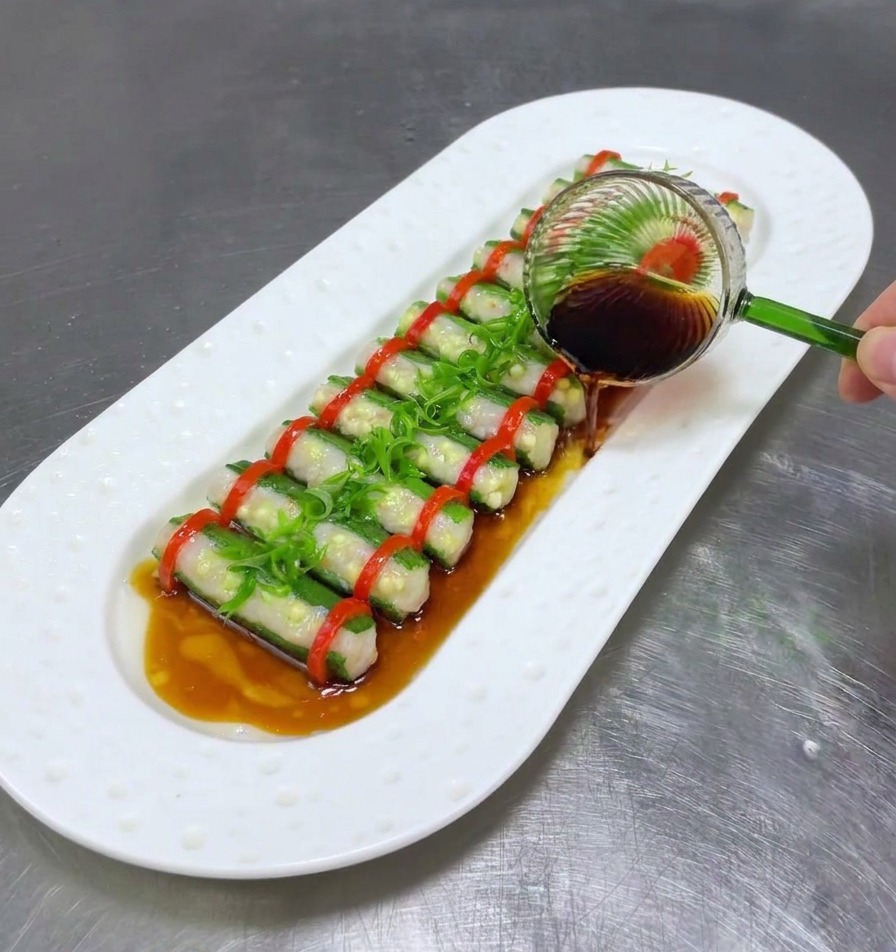 A glass ladle drizzling dark soy sauce over steamed stuffed okra and shredded scallions on a white plate.