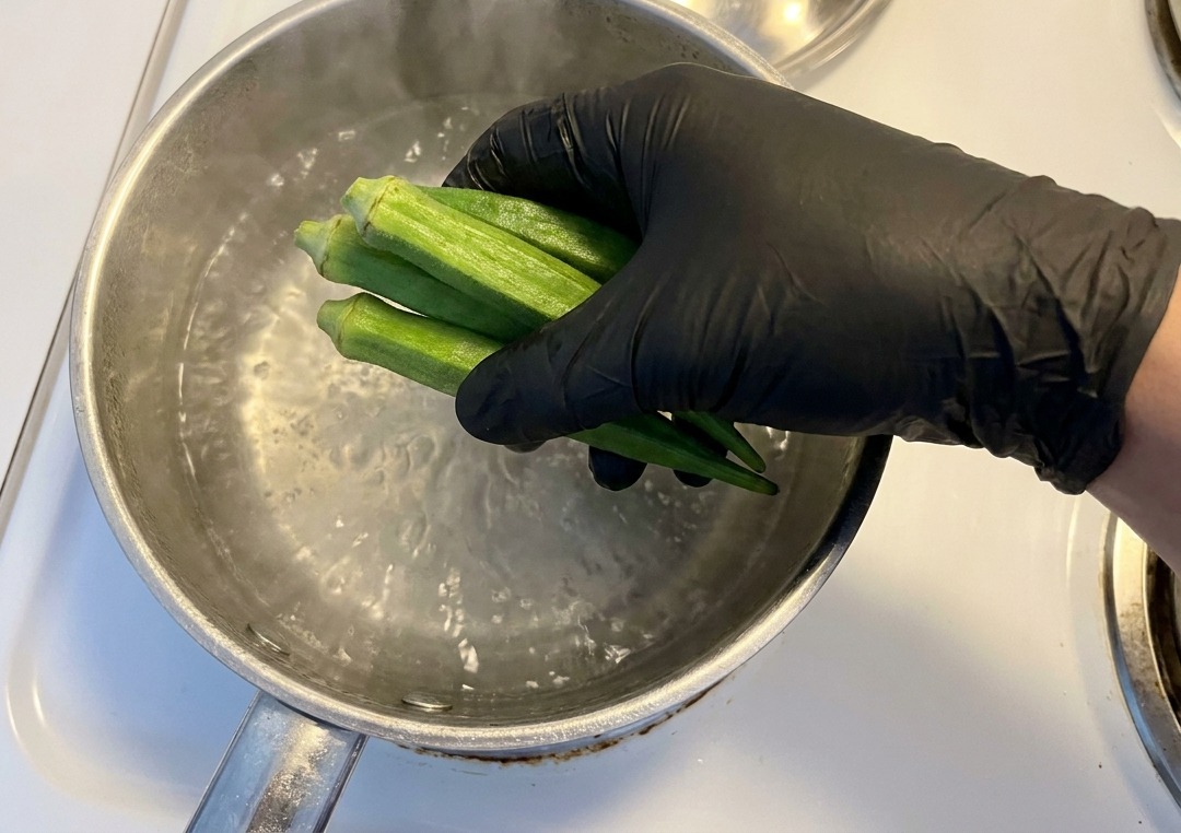 A gloved hand placing fresh, whole green okra pods into a stainless steel pot of boiling water on a white stove.