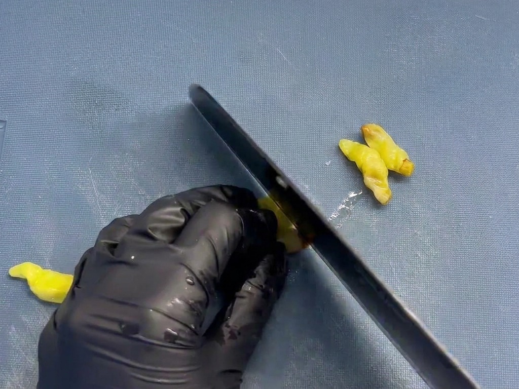 A chef using a black knife to finely mince small yellow pickled chili peppers on a blue textured cutting board.