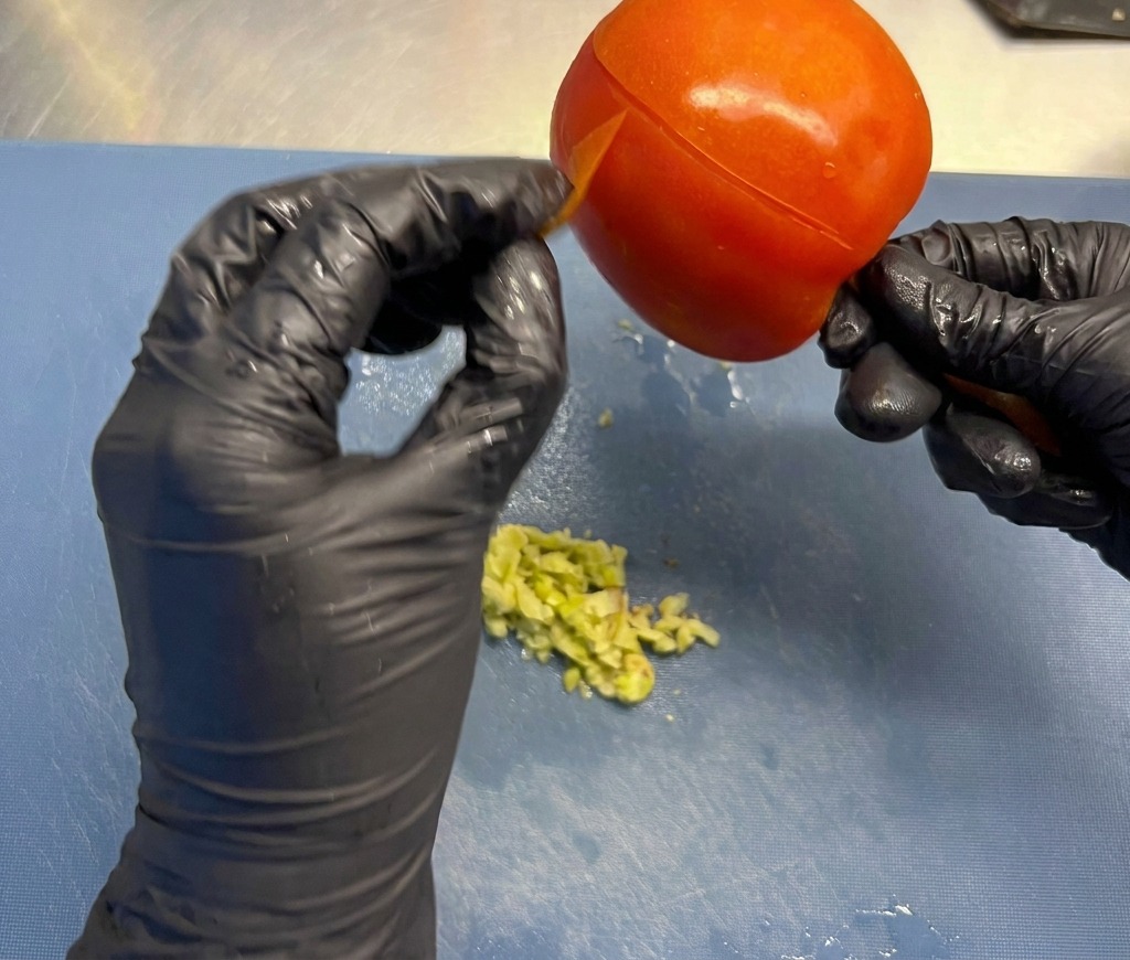 Hands in black gloves peeling the thin outer skin from a large, ripe red tomato over a preparation board.