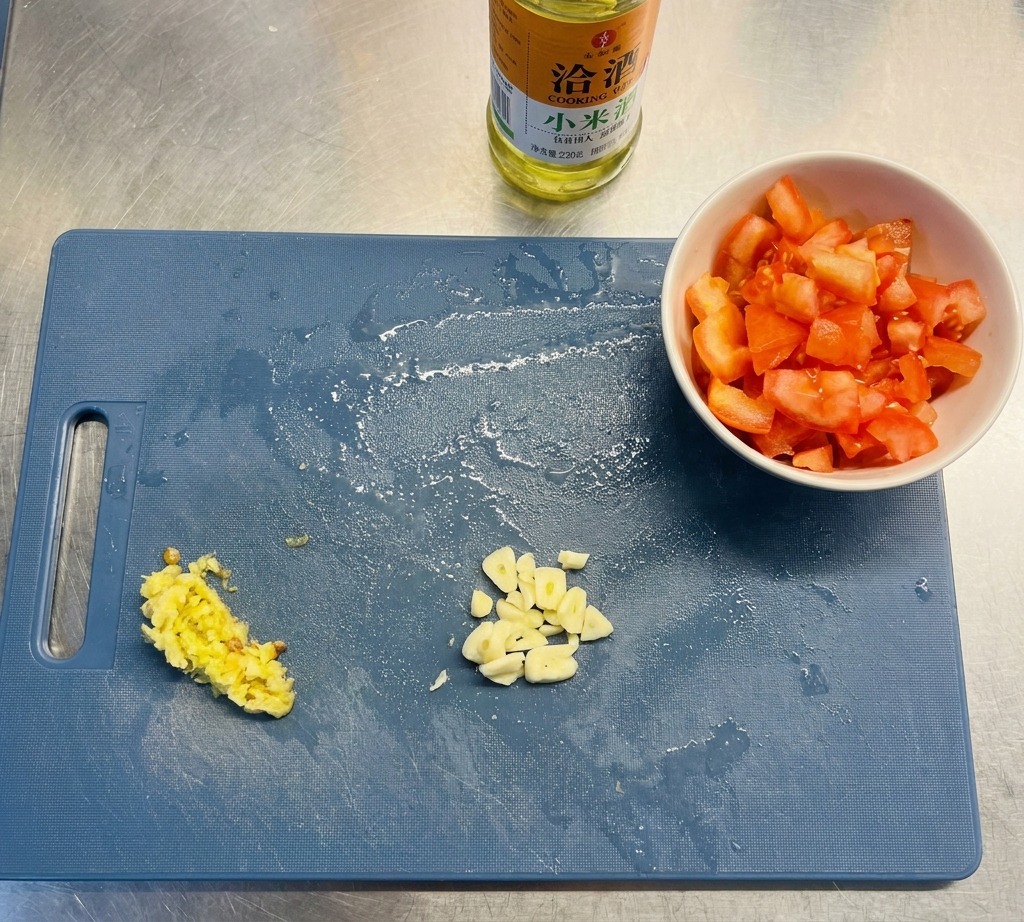 A blue cutting board showing prepared piles of sliced garlic, chopped yellow peppers, and diced tomatoes next to a bottle of oil.
