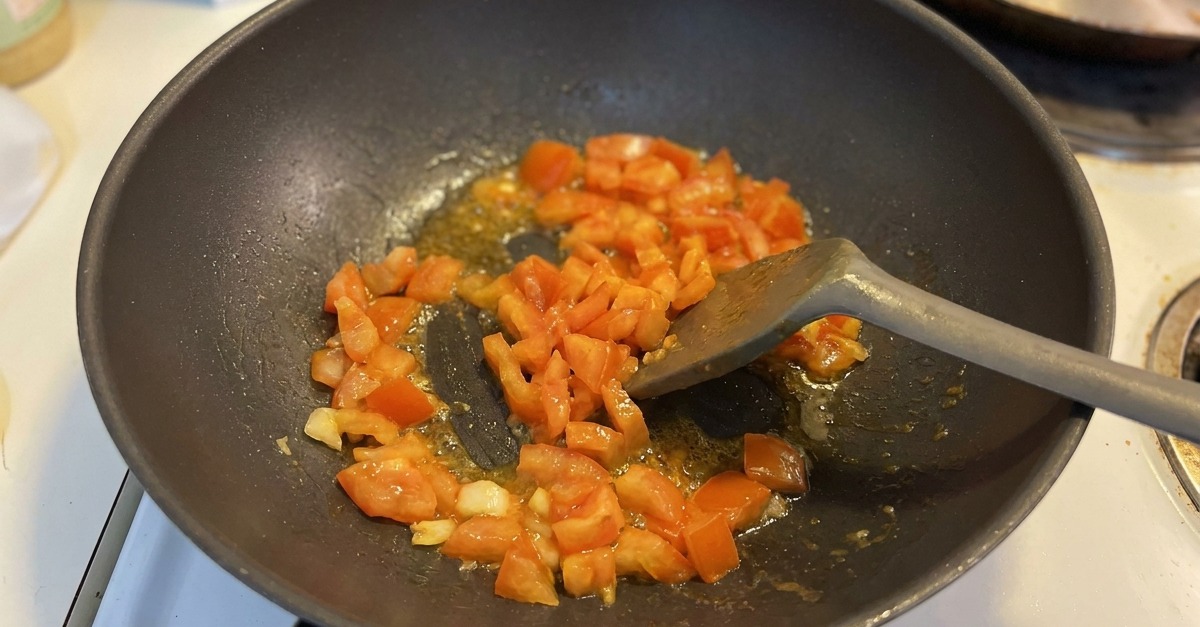 Diced tomato chunks being sautéed with garlic in a dark wok, with juices beginning to bubble at the bottom.