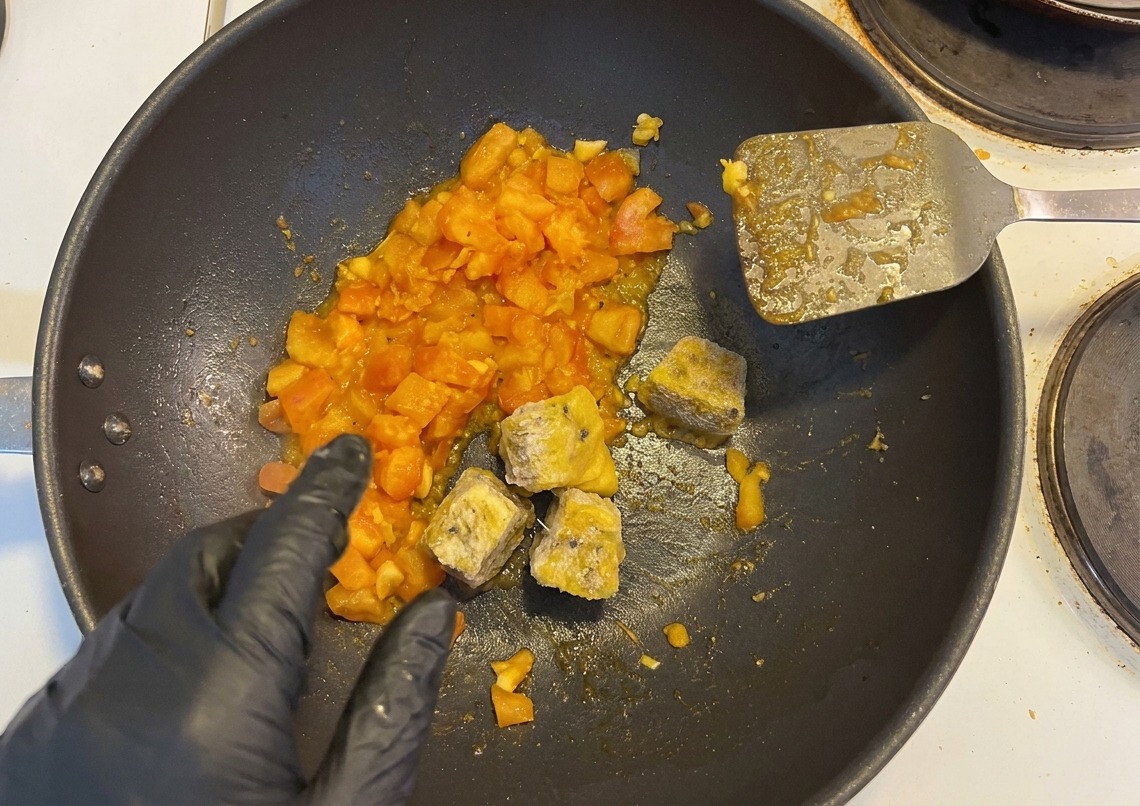 Frozen cubes of passion fruit pulp and diced pickled peppers being added to a pan of sautéed tomatoes by a hand in a black glove.