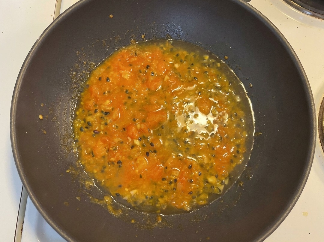 Water added to the tomato and passion fruit mixture in a dark wok, creating a vibrant orange soup base with visible passion fruit seeds.