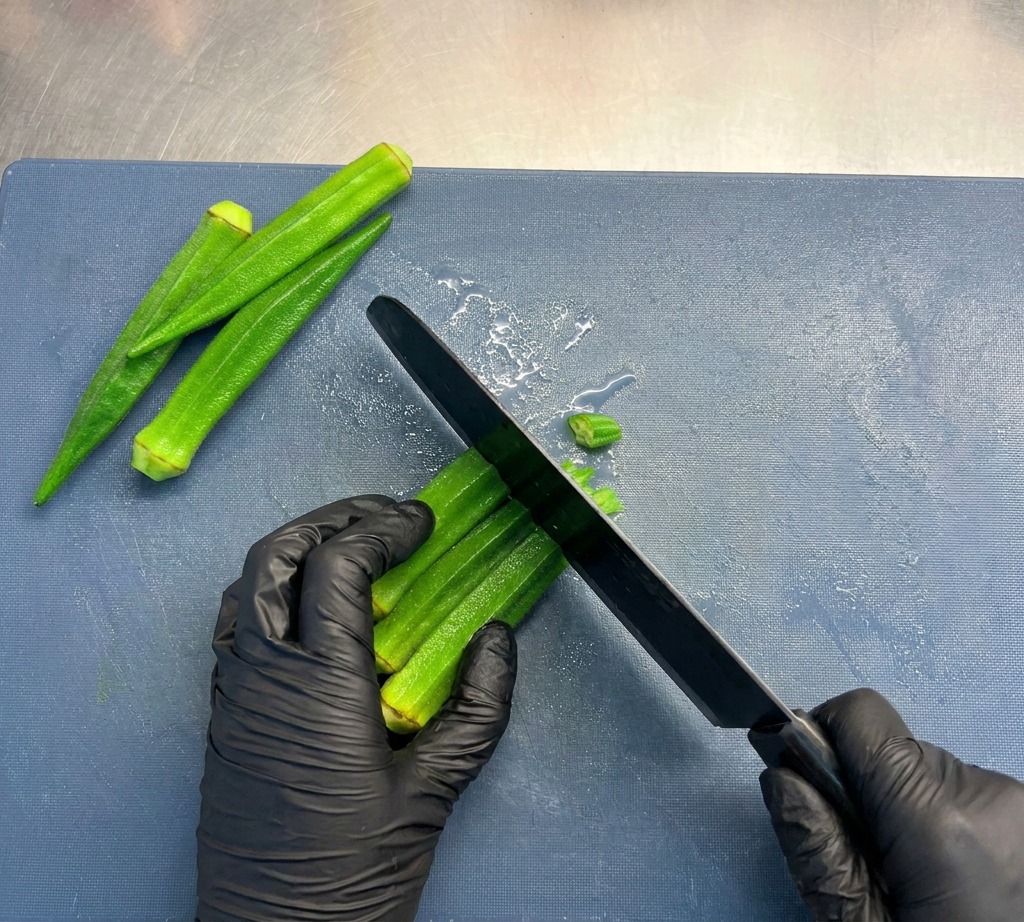 A person wearing black gloves using a chefs knife to slice bright green okra stalks into small circular rounds on a blue cutting board.