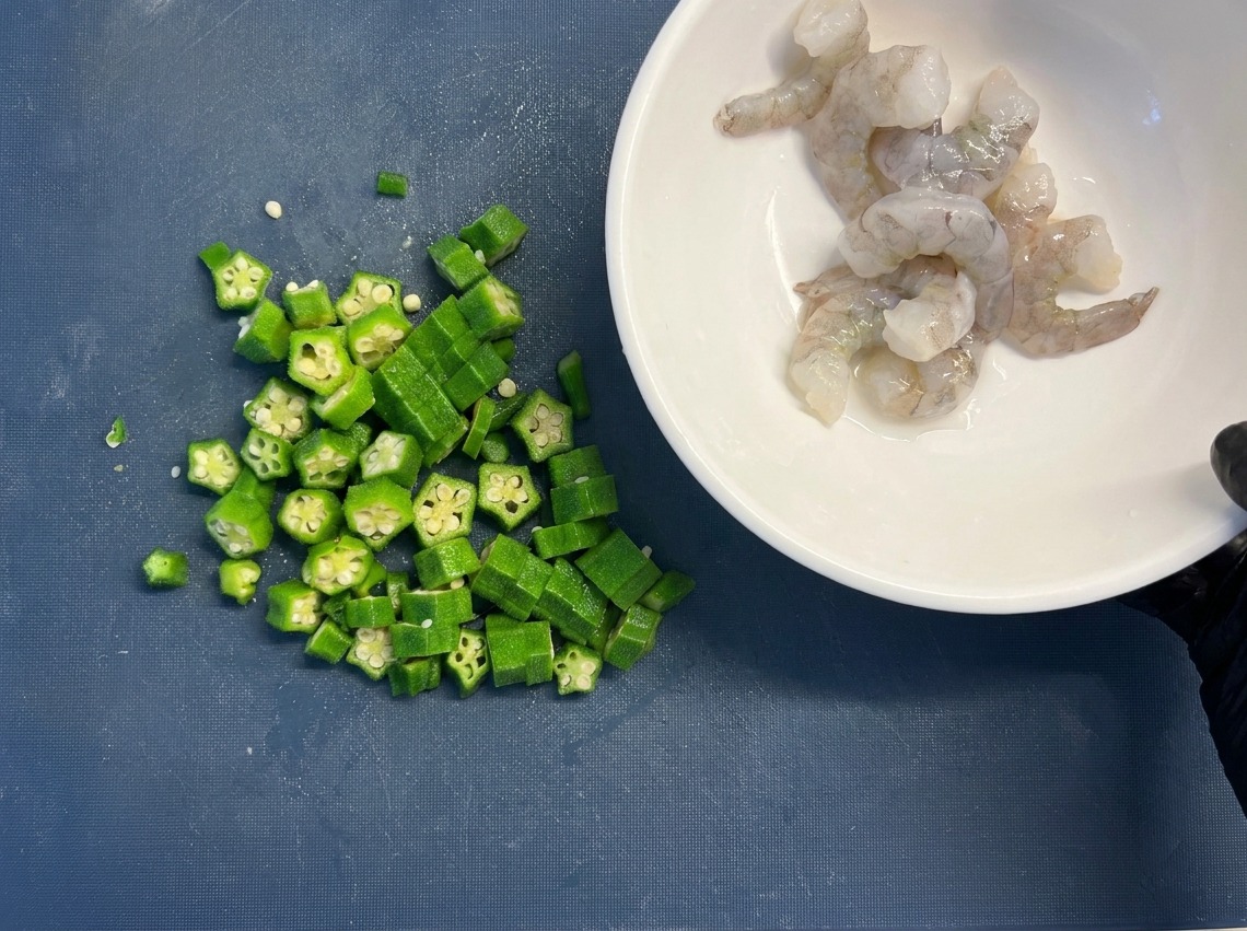 A white bowl filled with raw, cleaned shrimp held next to a pile of sliced green okra rounds on a blue cutting board.