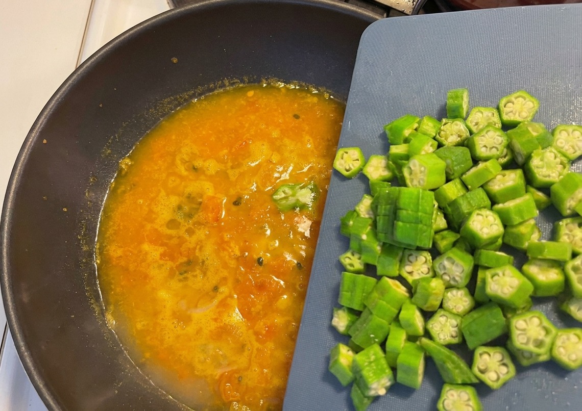 Freshly sliced okra rounds being slid from a blue cutting board into a bubbling orange soup in a dark wok.