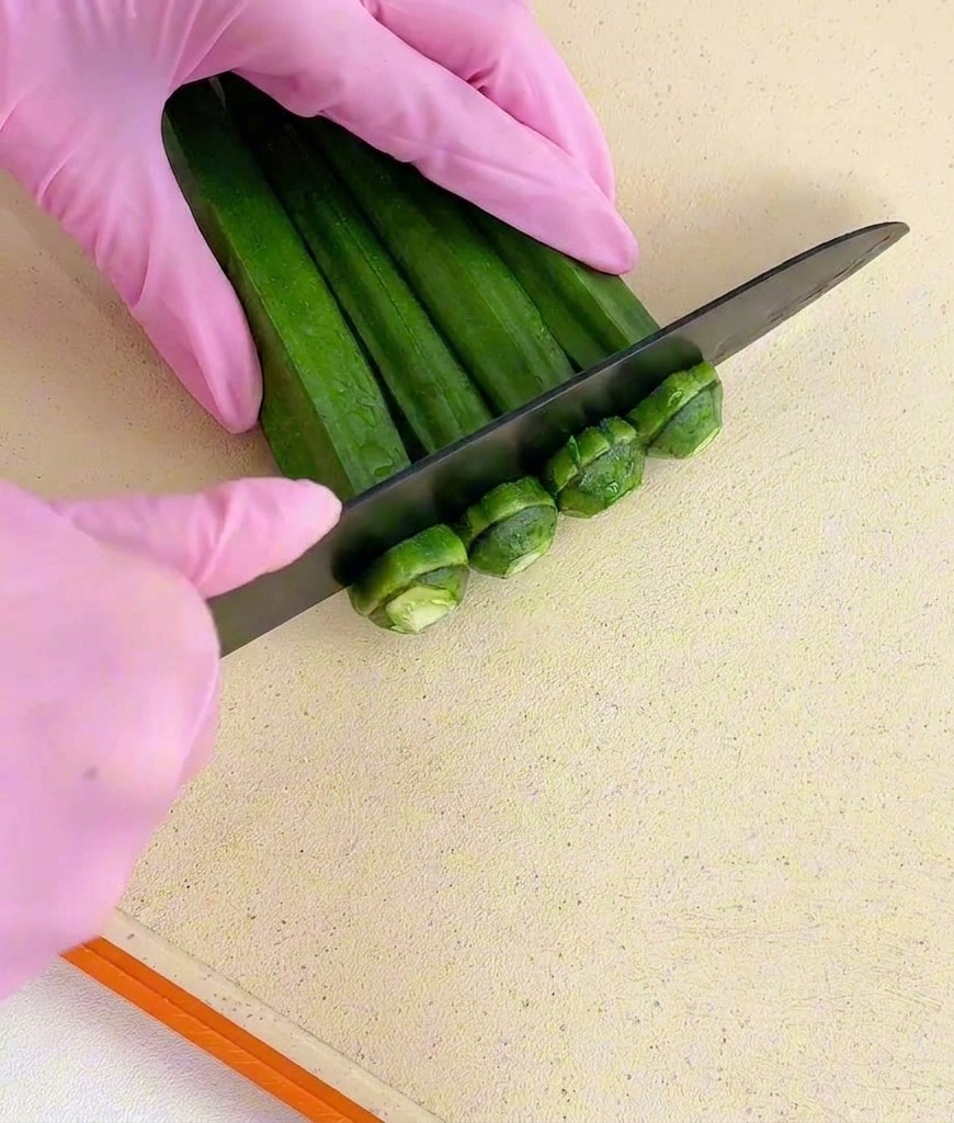Hands in pink gloves using a black knife to slice the stem ends off a bundle of green okra.