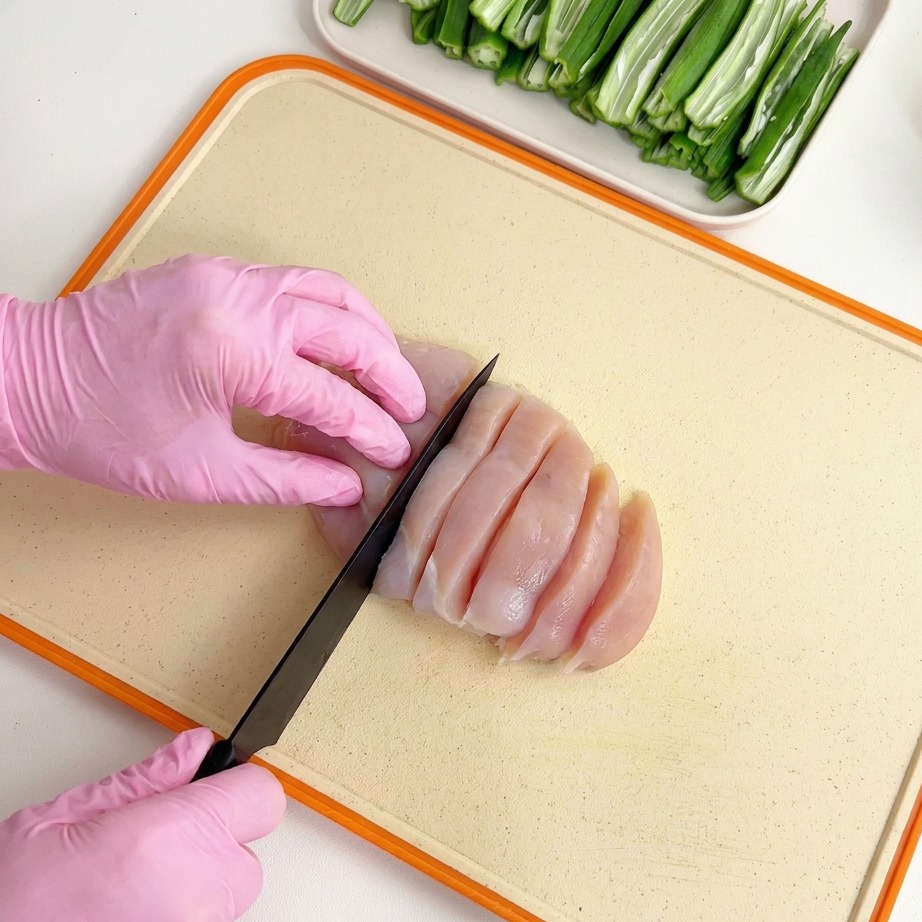 Raw chicken breast being sliced with a black knife on a light-colored cutting board by hands wearing pink gloves.