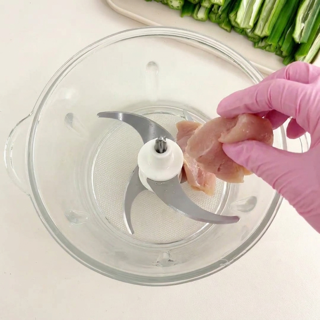A hand in a pink glove putting raw chicken chunks into a glass food processor container.