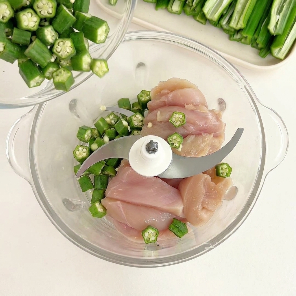 Green okra seeds being poured from a small bowl onto raw chicken pieces in a glass food processor.