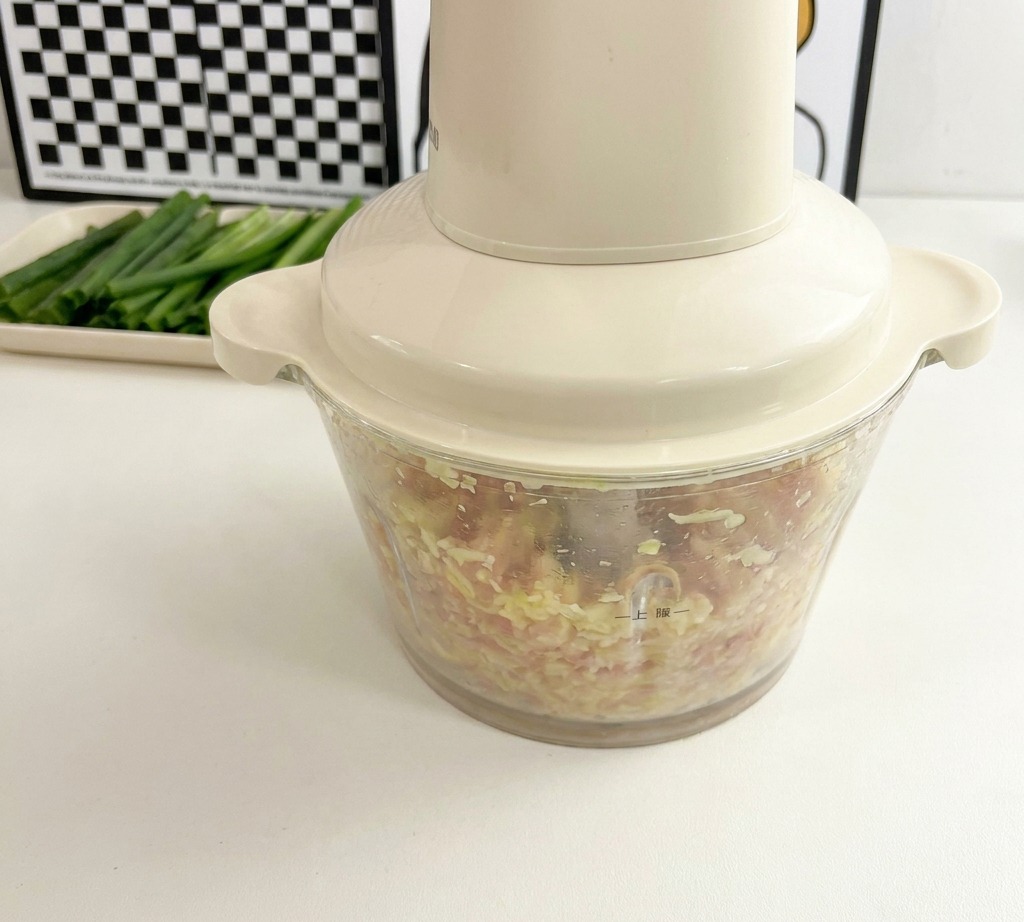 A cream-colored food processor sits on a white countertop, filled with a finely minced mixture of pink chicken meat and small yellowish okra seeds.