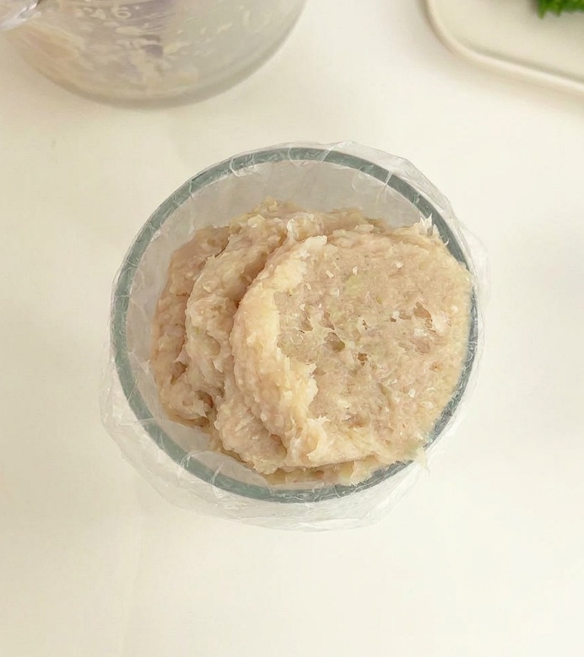 A clear bowl containing a light-colored, smooth meat paste lined with a plastic piping bag ready for use.