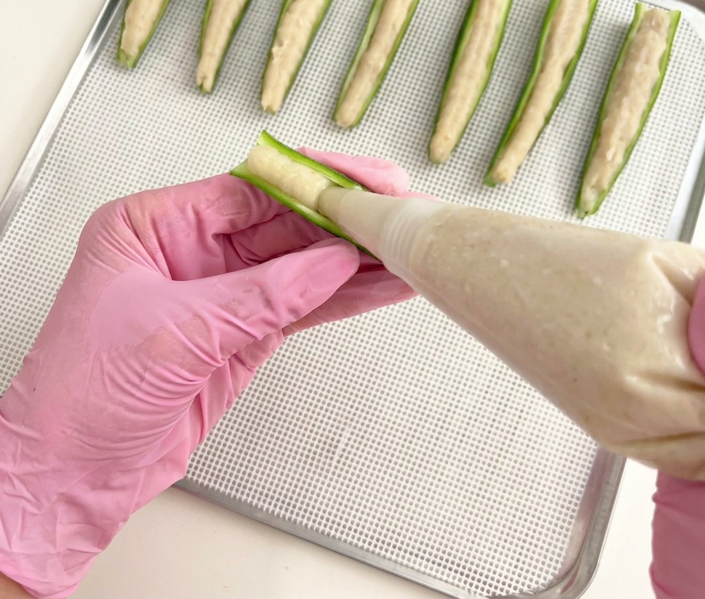 A person wearing pink gloves using a piping bag to fill green okra halves with a smooth chicken paste on a metal wire tray.