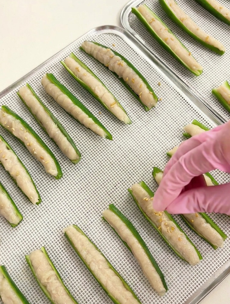 Close-up of a hand sprinkling white sesame seeds onto rows of okra stuffed with meat paste on a silver tray.