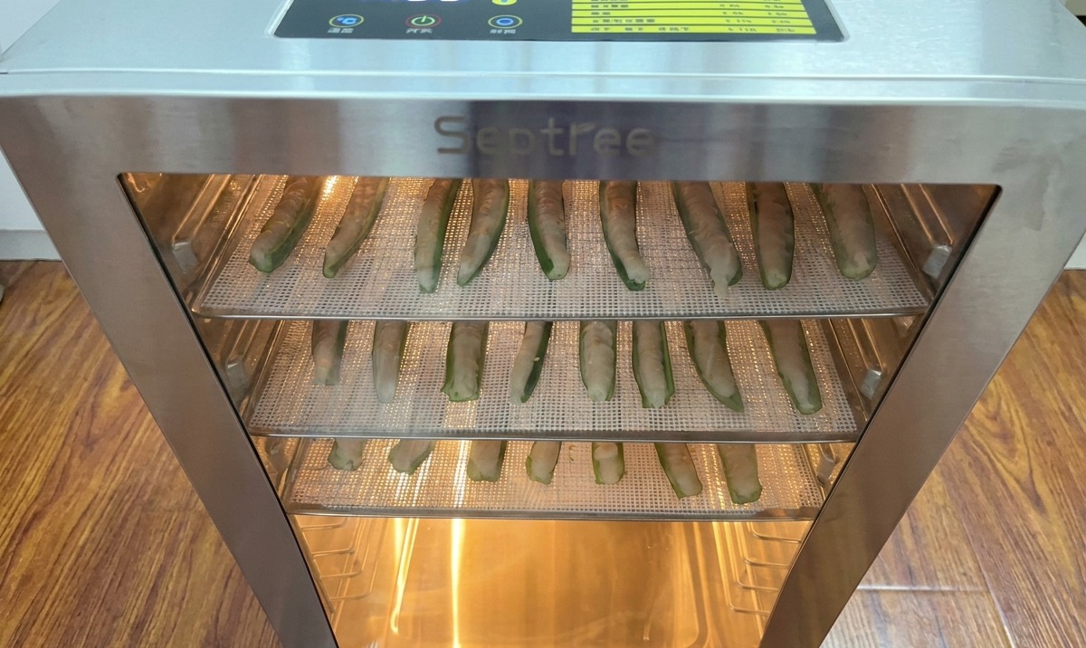Racks of okra halves filled with minced chicken meat being placed inside a stainless steel food dehydrator with a digital timer.