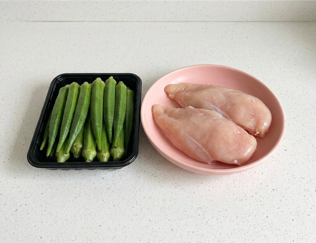 A black tray of fresh green okra standing vertically next to a pink plate containing two raw chicken breast fillets on a white countertop.
