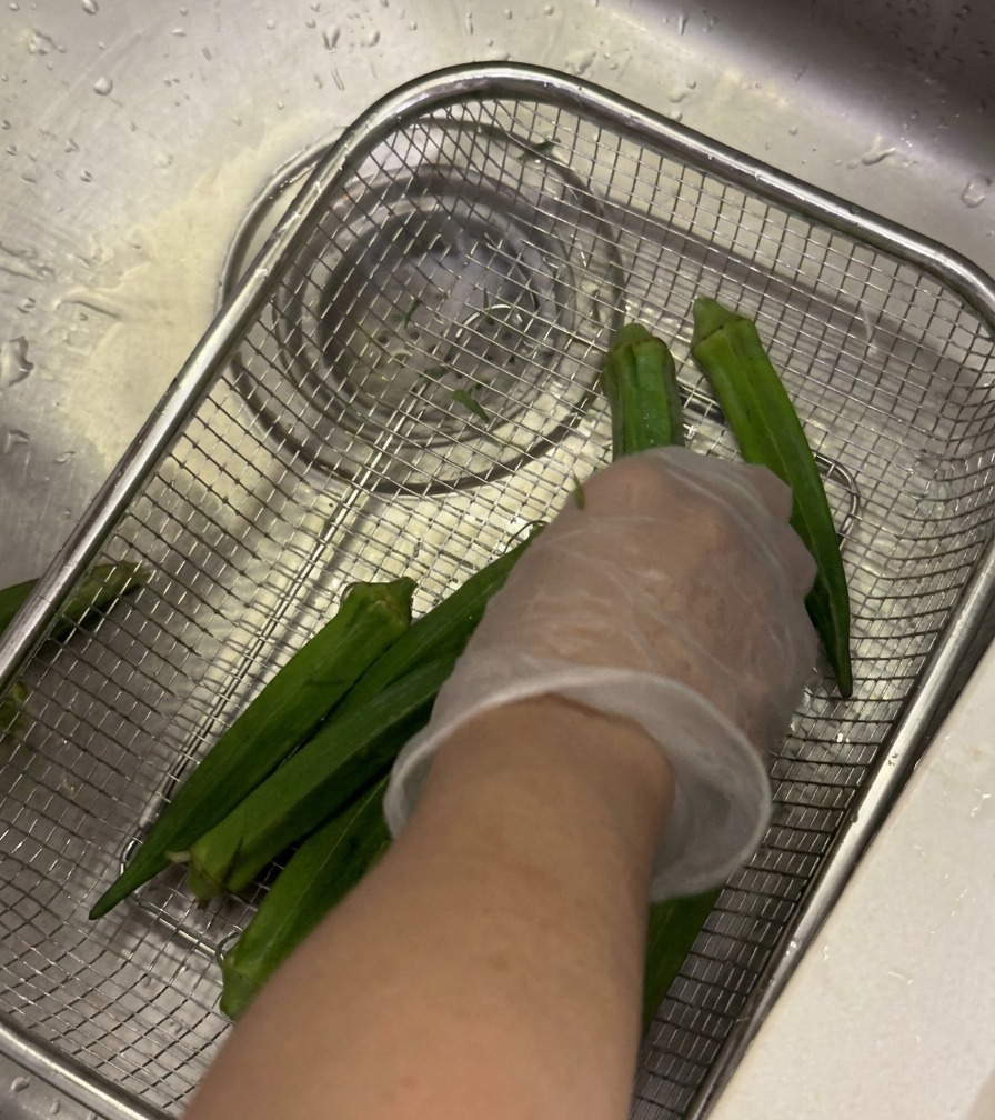 Fresh okra pods being washed in a wire mesh basket under a kitchen faucet with a gloved hand rubbing the pods.