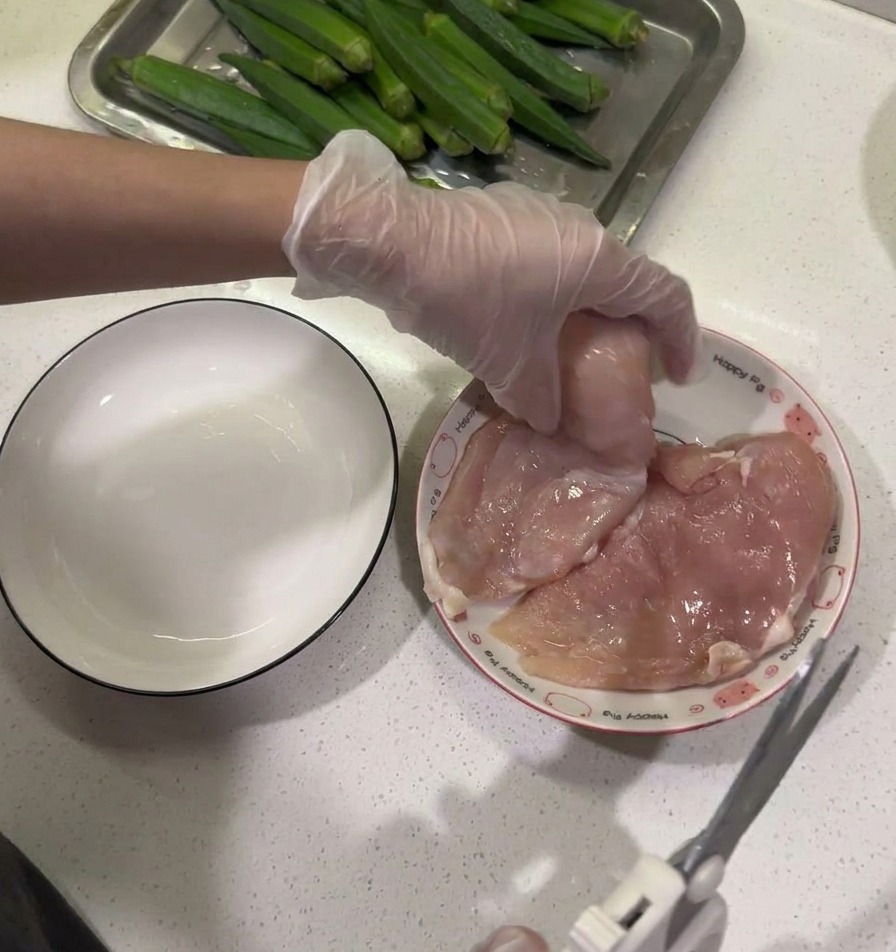 Kitchen shears being used to trim fat from a raw chicken breast held over a plate next to an empty bowl.