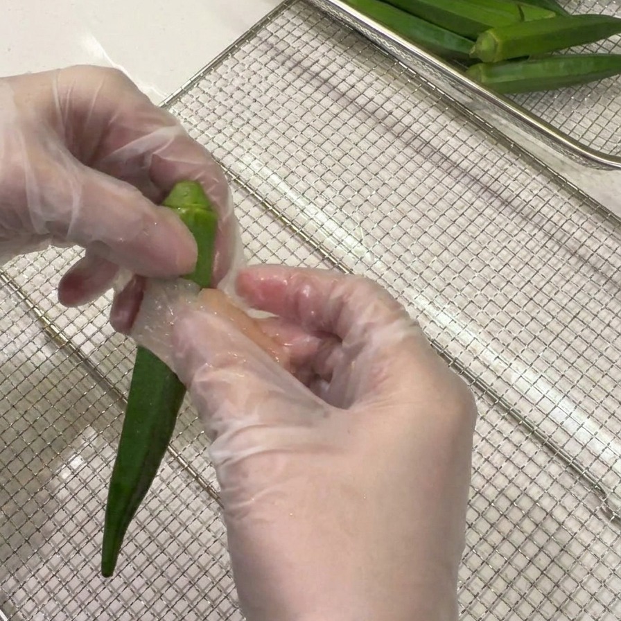 Gloved hands wrapping a thin slice of raw chicken breast in a spiral pattern around a whole green okra pod.
