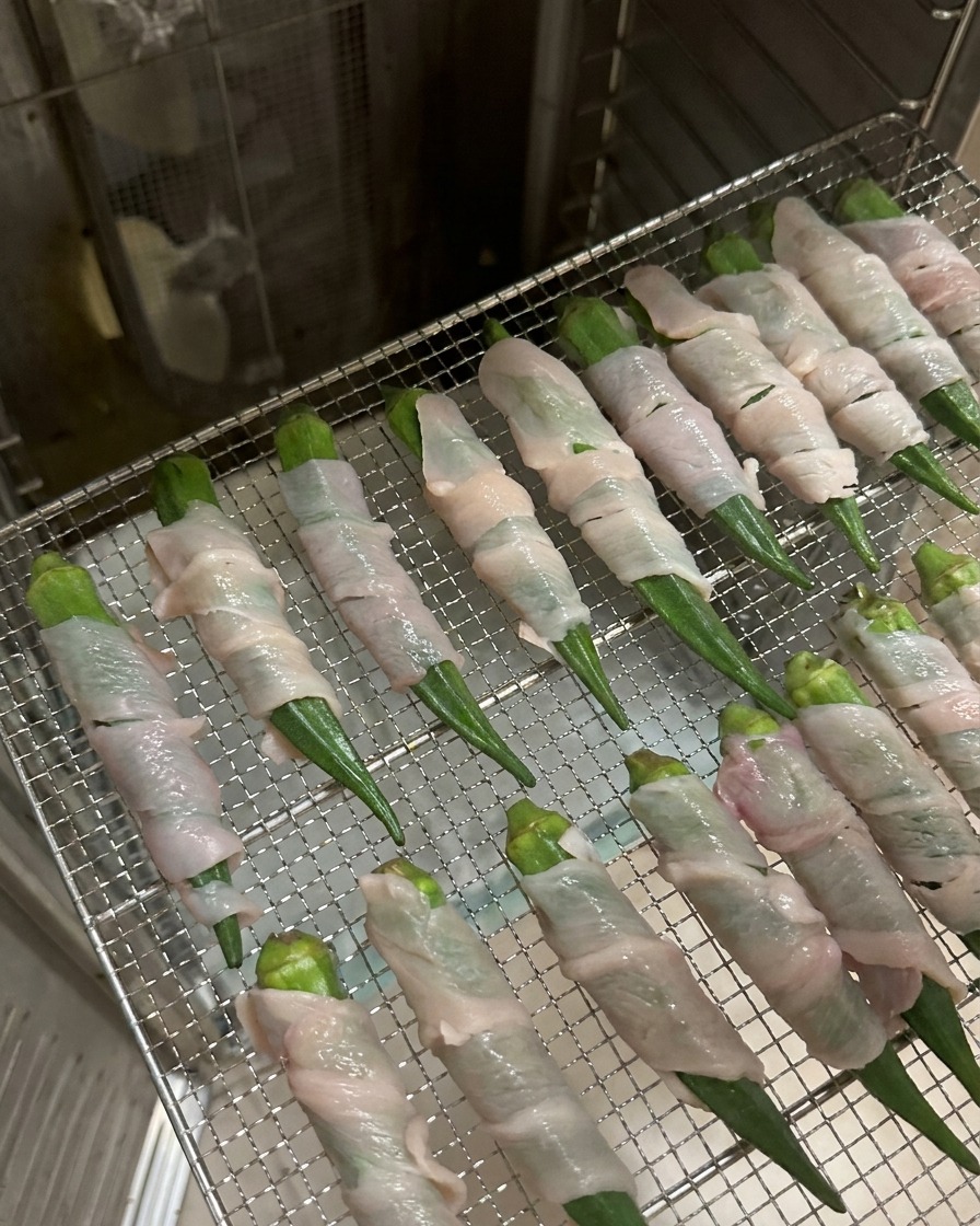 Rows of chicken-wrapped okra pods arranged on a metal wire rack inside a stainless steel dehydrator.