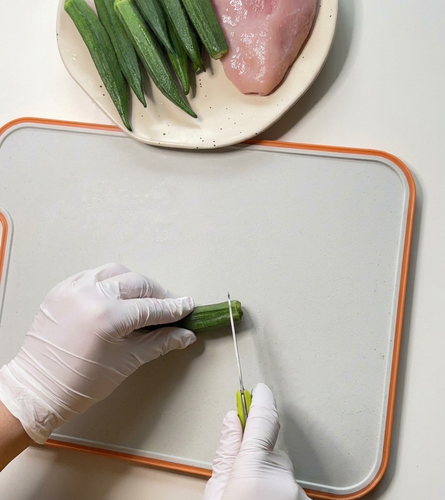 Gloved hands using a small paring knife to remove the ends of fresh green okra pods on a white cutting board.