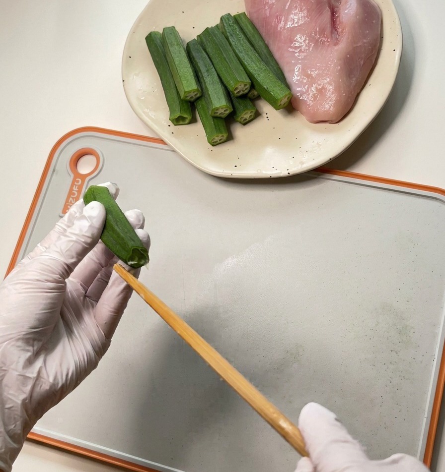 A person wearing white gloves uses a wooden chopstick to extract seeds and pulp from a green okra pod.