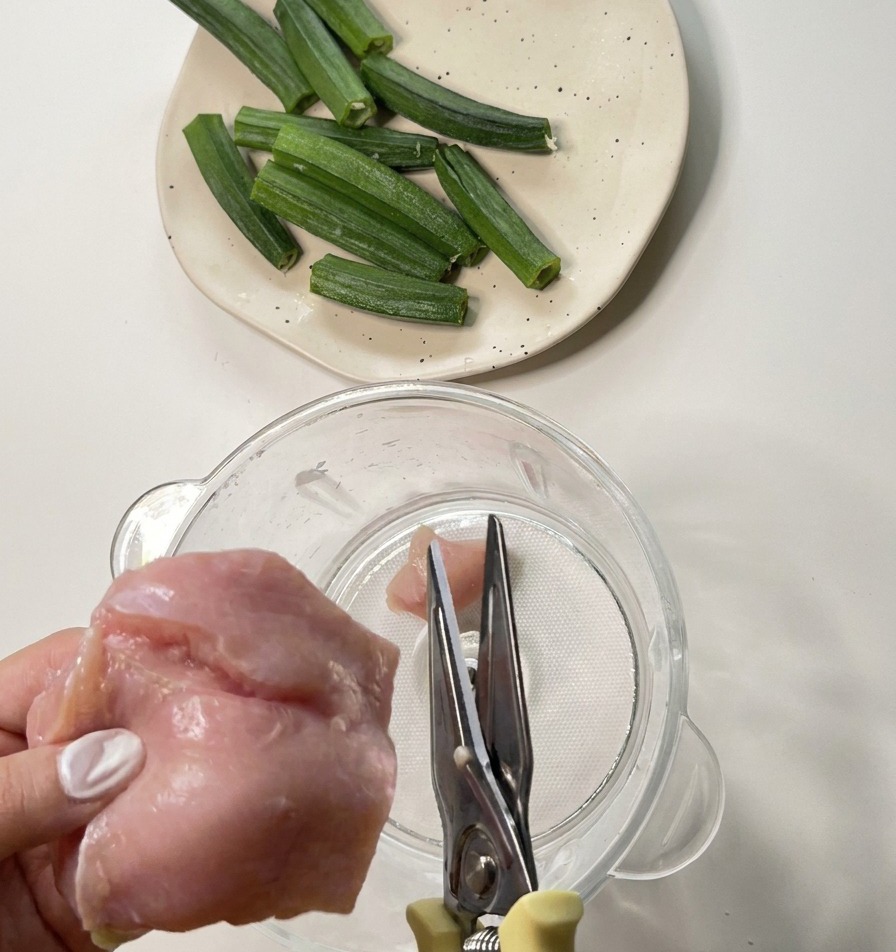 Hands using metal kitchen shears to cut raw chicken breast into a clear blender jar.