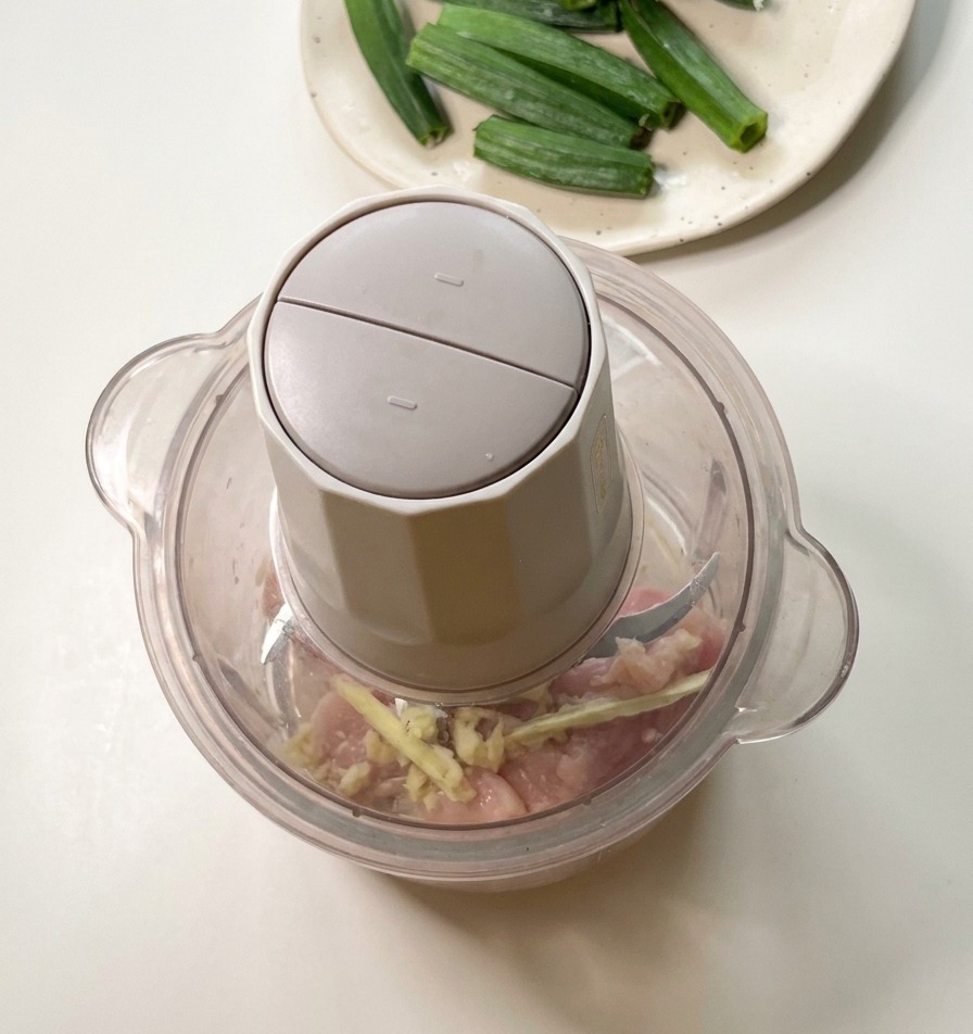 A food processor on a kitchen counter containing raw chicken pieces and okra seeds, ready for processing.