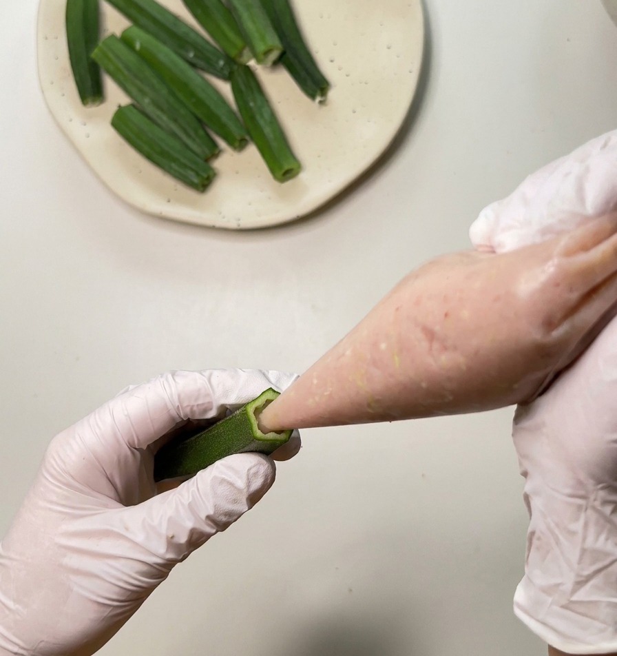 A close-up of a person piping pink chicken paste into a hollow green okra pod.