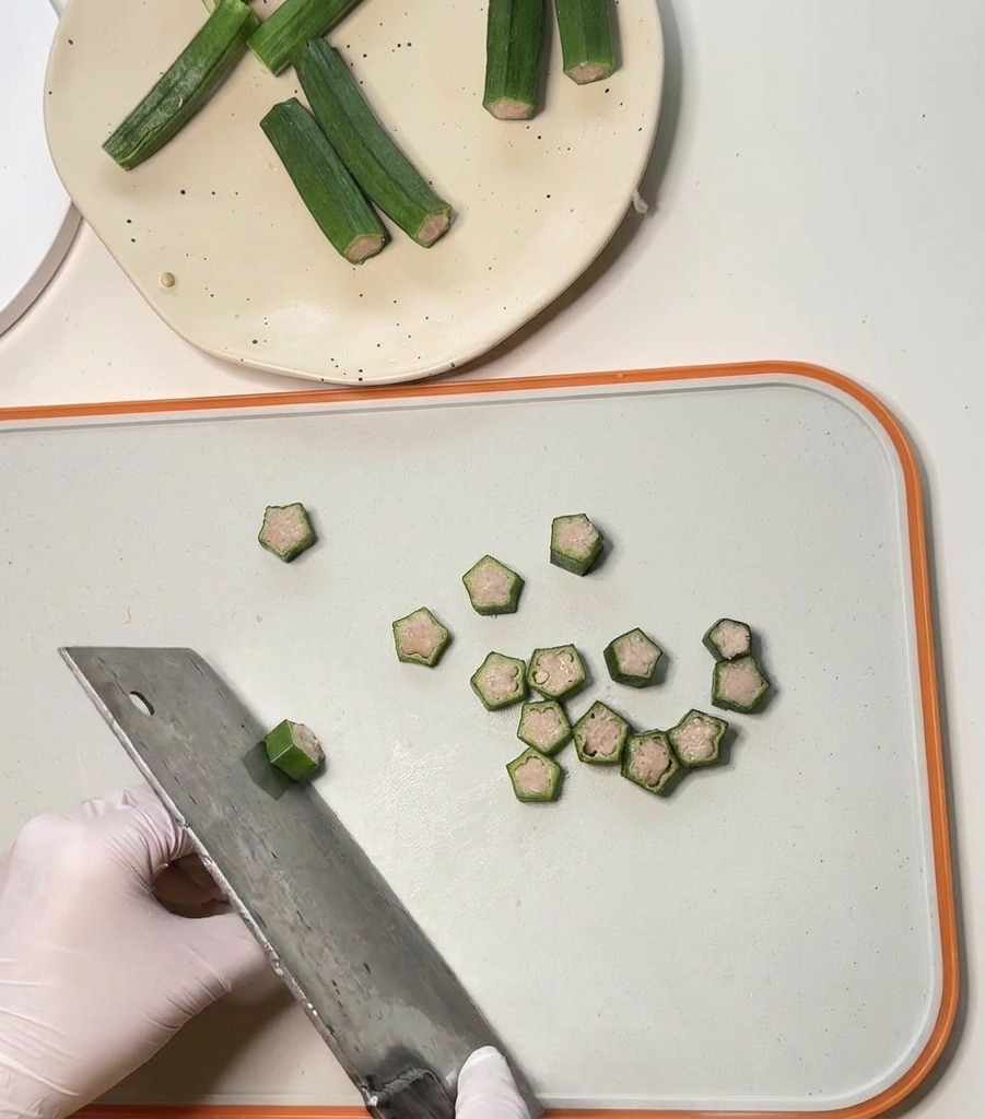A large metal cleaver slicing through chicken-stuffed okra pods into small, star-shaped pet treat rounds.