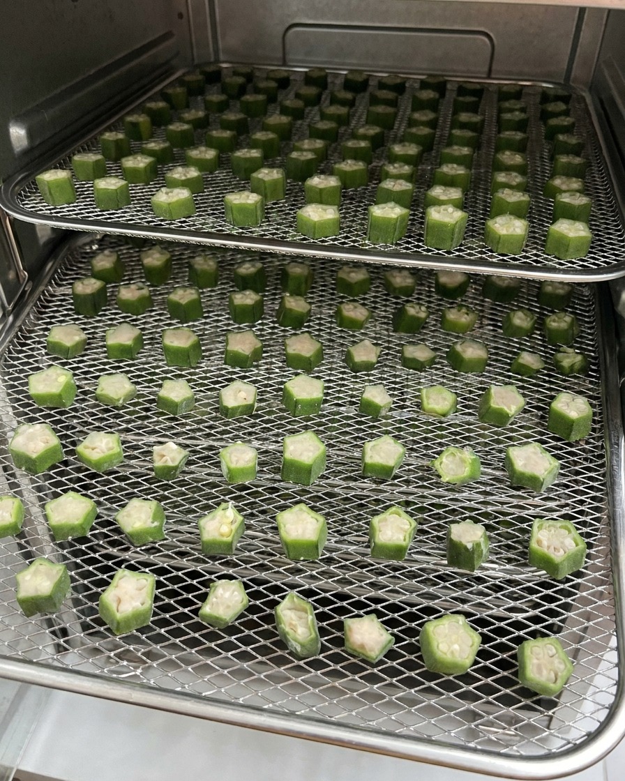 Two wire racks filled with sliced okra and chicken pet treats sitting inside a food dehydrator during the drying process.