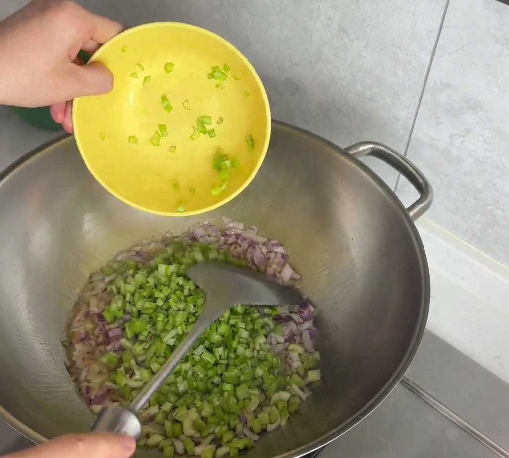 A person pouring diced celery from a yellow bowl into a stainless steel pan containing chopped red onions.