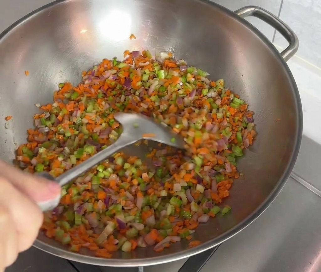 A colorful mixture of finely diced carrots, celery, and red onions being stir-fried with a spatula in a stainless steel wok.