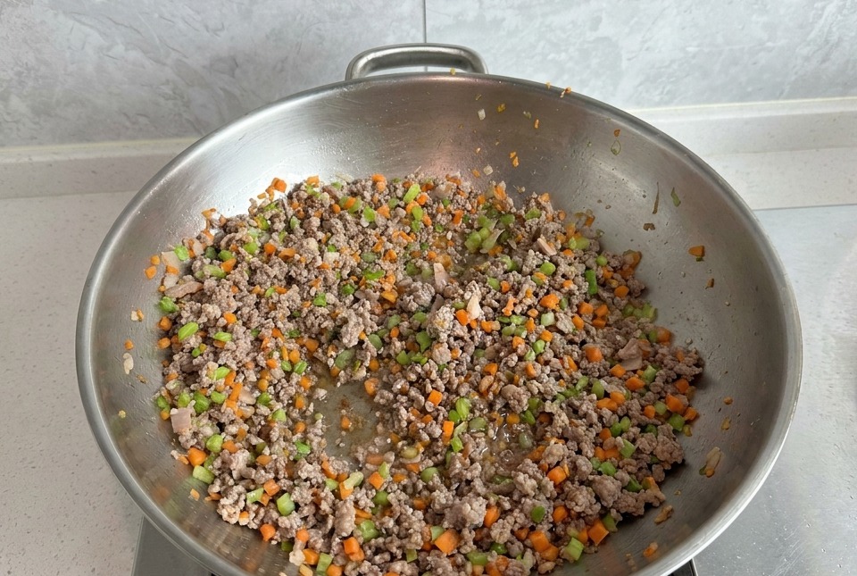 Minced meat being stir-fried with diced carrots and celery in a large silver wok, with the meat appearing mostly browned.